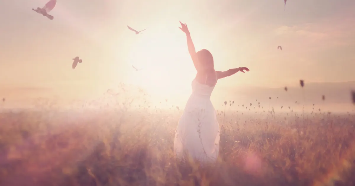 Woman in a white dress with arms raised in a field at sunrise